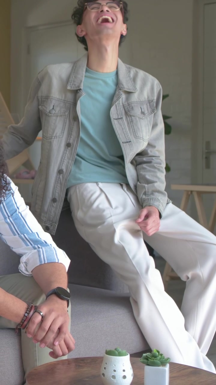 Vertikal video: Smilende mand i denimjakke griner, efter en ven har talt, derhjemme med sukkulenter.