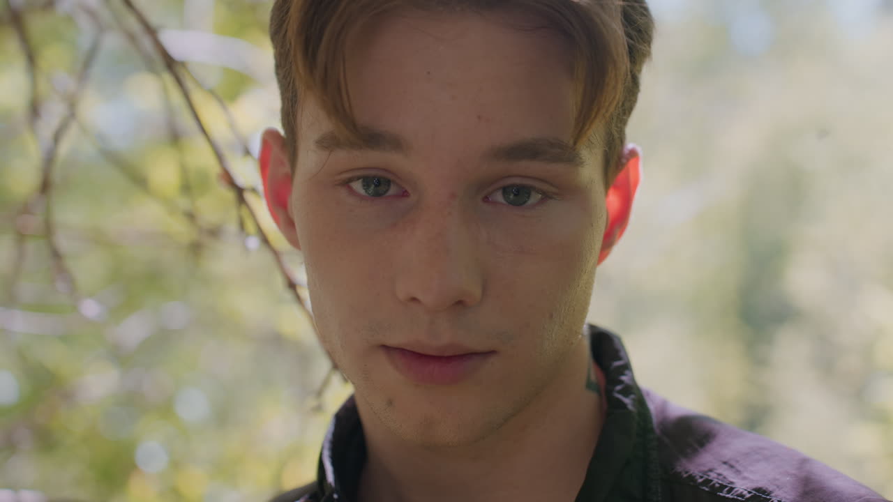 Close-view of weary young male tourist standing in forest, eyes shut and hands pressed against nose in gesture of exhaustion, while a subtle expression of hope flickers across tired face