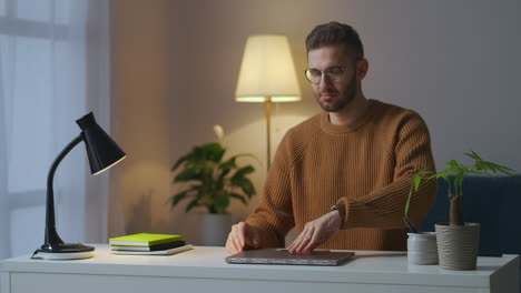 Un-Programador-Masculino-Está-Abriendo-Un-Cuaderno-Para-Trabajar-En-Casa-Por-La-Noche-Encendiendo-Una-Lámpara-De-Mesa,-Un-Especialista-Independiente-En-Trabajo-Remoto