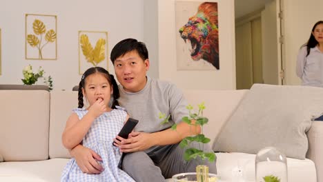 father and daughter enjoy tv time on a couch, with a woman entering the room. bright, warm lighting enhances the cozy atmosphere