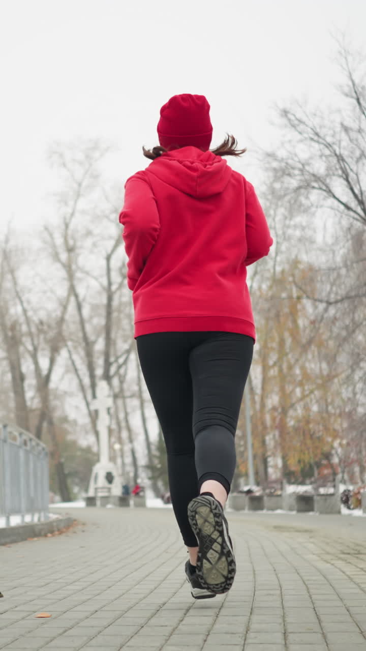 back view of athlete jogging along snowy park path wearing red hoodie and black leggings near iron railing surrounded by winter scenery with white building and trees showing cold fitness motion