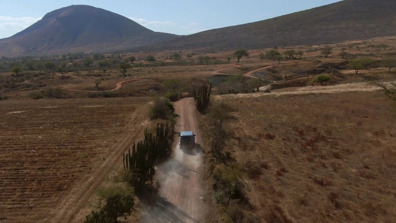 Vista aérea do campo com veículo a percorrer uma estrada rural empoeirada através de campos secos e terreno montanhoso acidentado sob um céu diurno claro