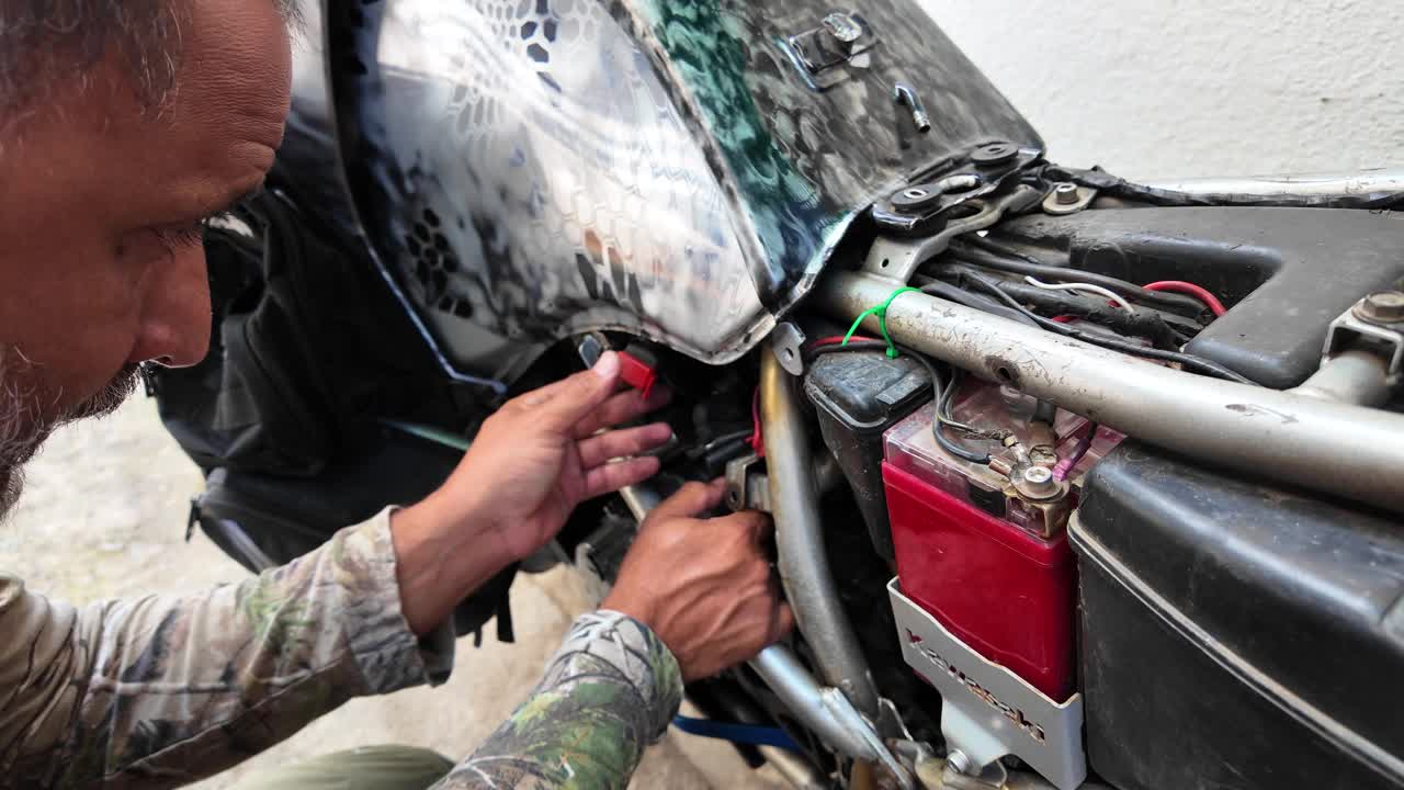 Focused mechanic troubleshooting the electrical system and ignition relay of a camouflage adventure motorcycle in a home workshop