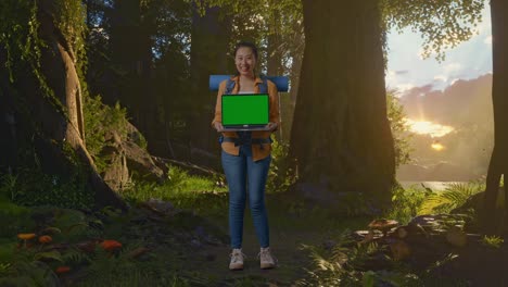cuerpo lleno de excursionista femenina asiática con mochila de montañismo sonriendo y mostrando simulacro de computadora portátil de pantalla verde mientras explora la naturaleza del bosque