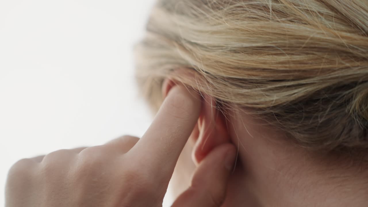 Primo piano di donna che si aggiusta l'orecchio con un dito, illuminazione clinica calma. Capelli biondi raccolti dietro l'orecchio, delicato massaggio con la punta del dito per ispezione uditiva e comfort. Focus sull'anatomia dell'orecchio e un sottile sollievo.