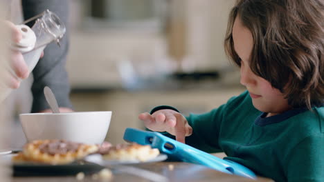 happy little boy using tablet computer at breakfast table in kitchen father pouring milk into cereal bowl enjoying caring for son at home