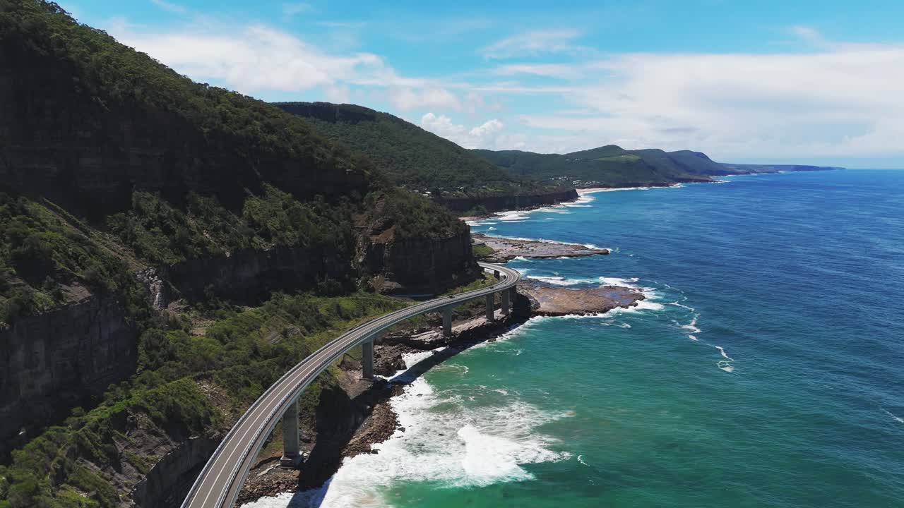 Backward-flying drone arcs left to unveil Australia’s Sea Cliff Bridge on Lawrence Hargrave Drive, tracking north toward Sydney and showcasing traffic sheer seaside cliffs and sparkling Pacific waters