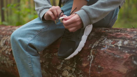 close-up of woman with long nails tying shoelaces on black sneakers while seated on fallen tree during hike, wearing gray sweater and light blue jeans