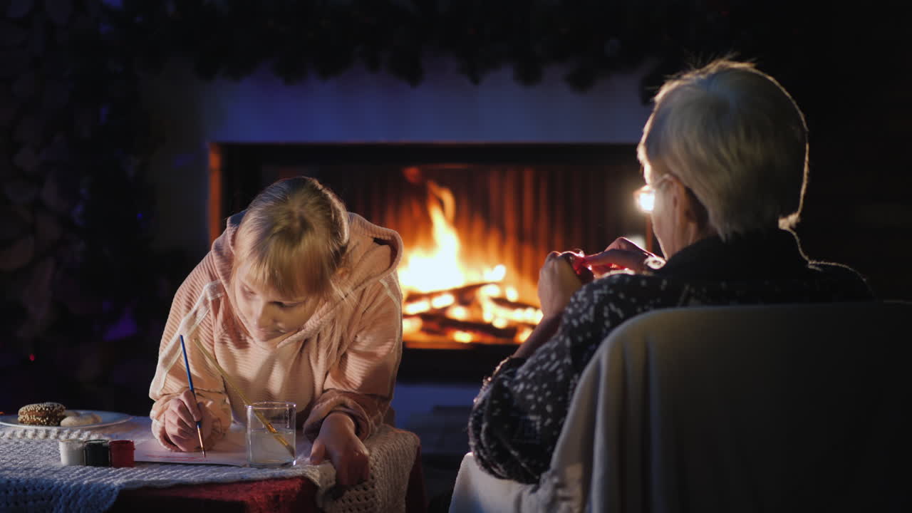 크리스마스 이브에 어린 소녀와 함께 있는 randmother는 벽난로 옆에서 휴식을 취합니다. 소녀는 편지를 씁니다.