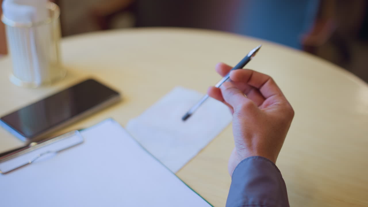 Close-up of man holding pen above blank paper on round table with smartphone and tissue in background, capturing moment of decision, pause, or thought before writing in quiet indoor setting