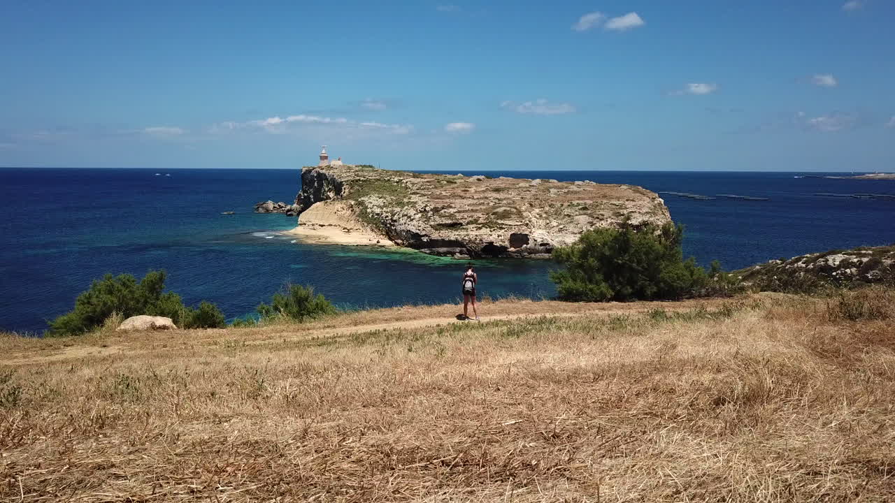 Drone shot of a woman standing infront of a beautiful view of a small, uninhabeted island in malta on a bright, sunny day.
