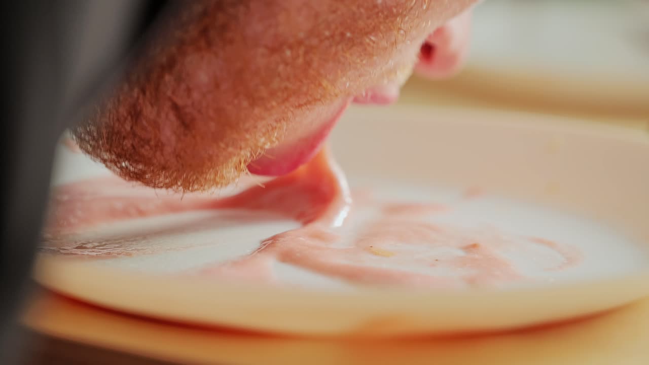 Red beard man slowly licks plate with pink banana strawberry sauce, slow motion