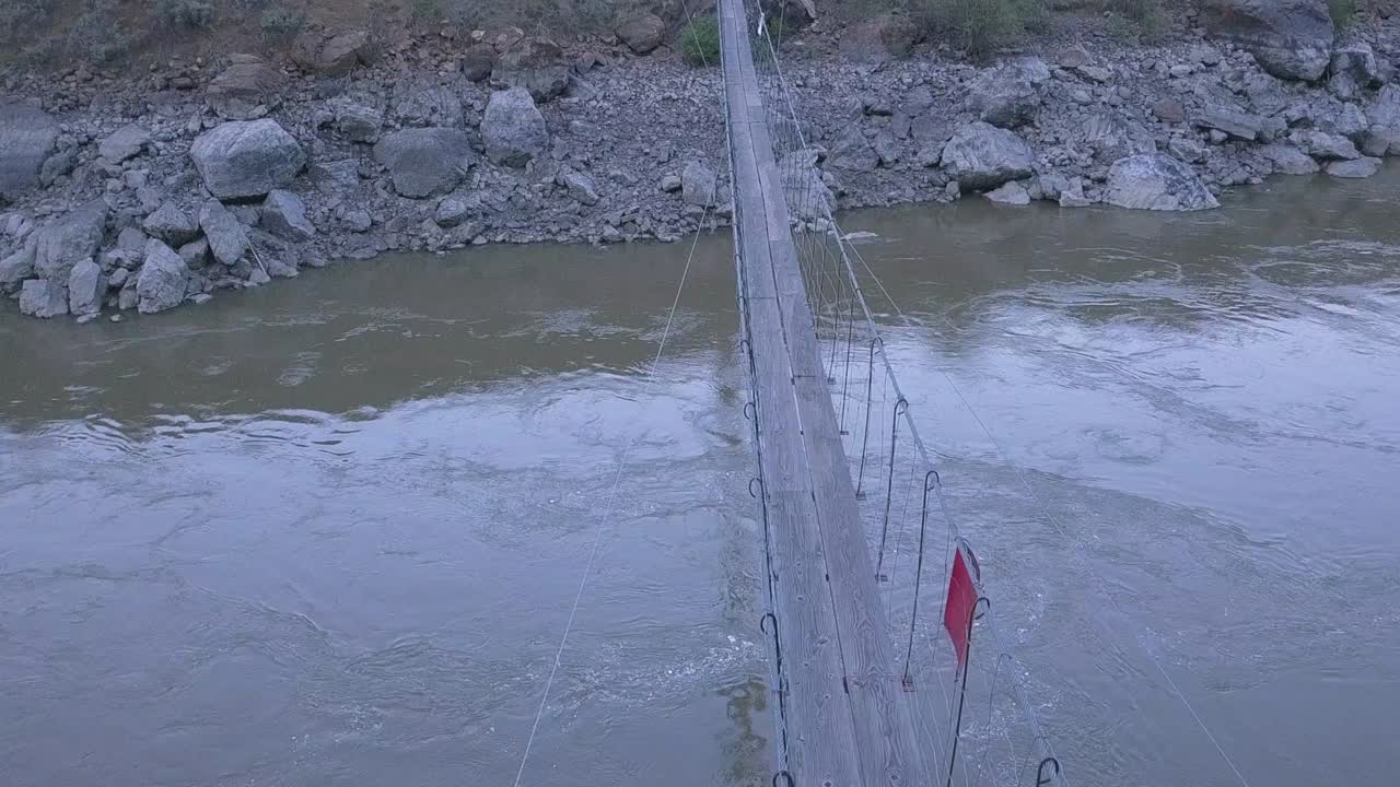 POV aerial: Narrow foot suspension bridge over muddy river canyon