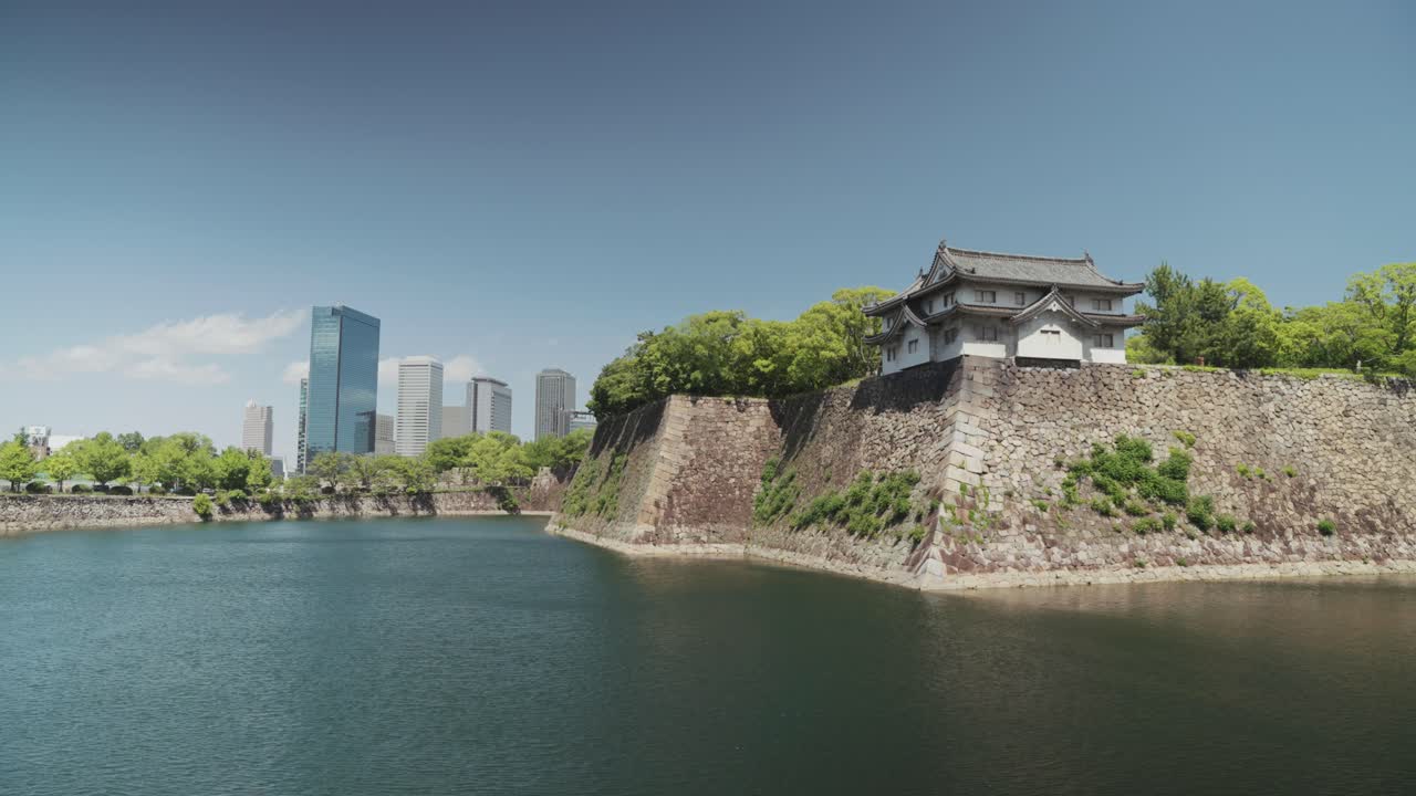Stunning view of Osaka Castle and its moat, surrounded by lush greenery, with modern skyscrapers in the background under a bright blue sky, blending history and urban scenery.
