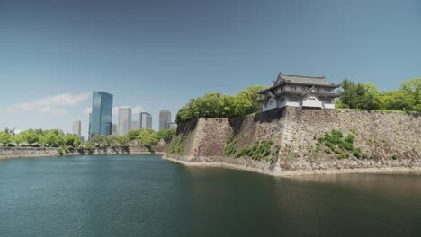stunning view of osaka castle and its moat, surrounded by lush greenery, with modern skyscrapers in the background under a bright blue sky, blending history and urban scenery.