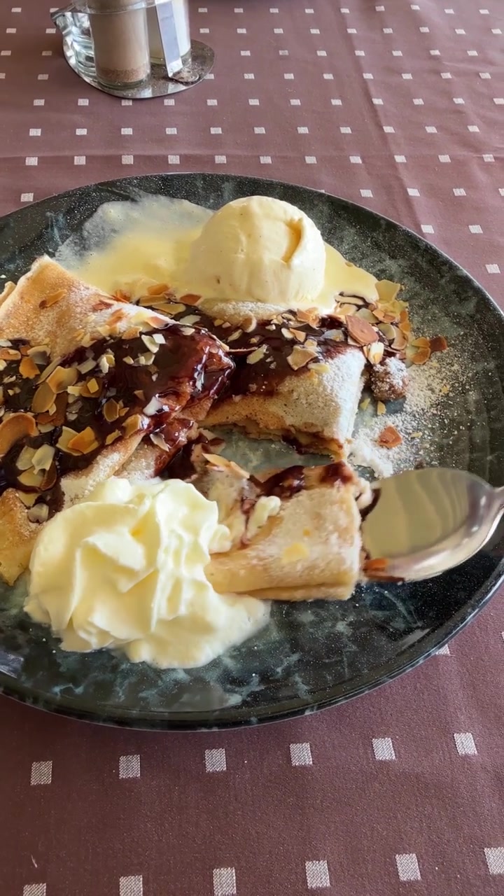 Dolce crepe guarnita con salsa al cioccolato, panna montata, gelato alla vaniglia, zucchero a velo e scaglie di mandorle, servita su un piatto in un'accogliente atmosfera da ristorante. Filmato verticale da iPhone. Vlog di cibo
