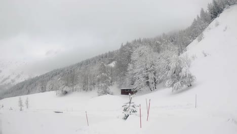 Scenic-Slow-Pan-of-Cabin-Amidst-Snowy-Mountainside,-Snow-Covered-Trees,-Light-Snowfall,-and-Cloudy-Sky