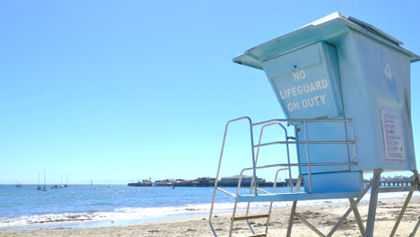 Sitzen-Am-Sandstrand-Und-Beobachten-Die-Blauen-Pazifischen-Ozeanwellen-Gegen-Die-Küste-Mit-Einem-Leeren-Rettungsturm-An-Einem-Sonnigen-Klaren-Tag-In-Santa-Barbara,-Kalifornien