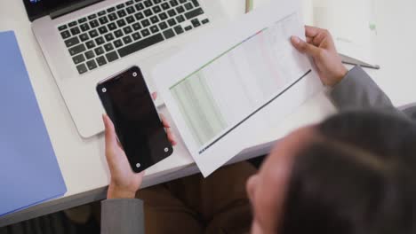 Mujer-De-Negocios-Birracial-Sentada-En-El-Escritorio,-Usando-Un-Teléfono-Inteligente-Con-Espacio-Para-Copiar-En-Una-Oficina-Moderna