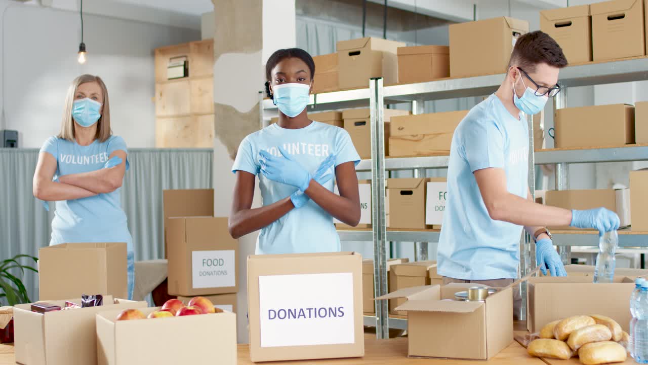 Multiethnic group of volunteers in facial mask packing boxes and looking at the camera in charity warehouse