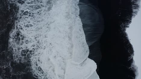 A-beautiful-drone-shot-of-the-Black-Beach-in-Iceland-shows-the-high-waves-of-the-sea-and-the-cloudy-weather-while-the-snow-covers-some-of-the-black-sand