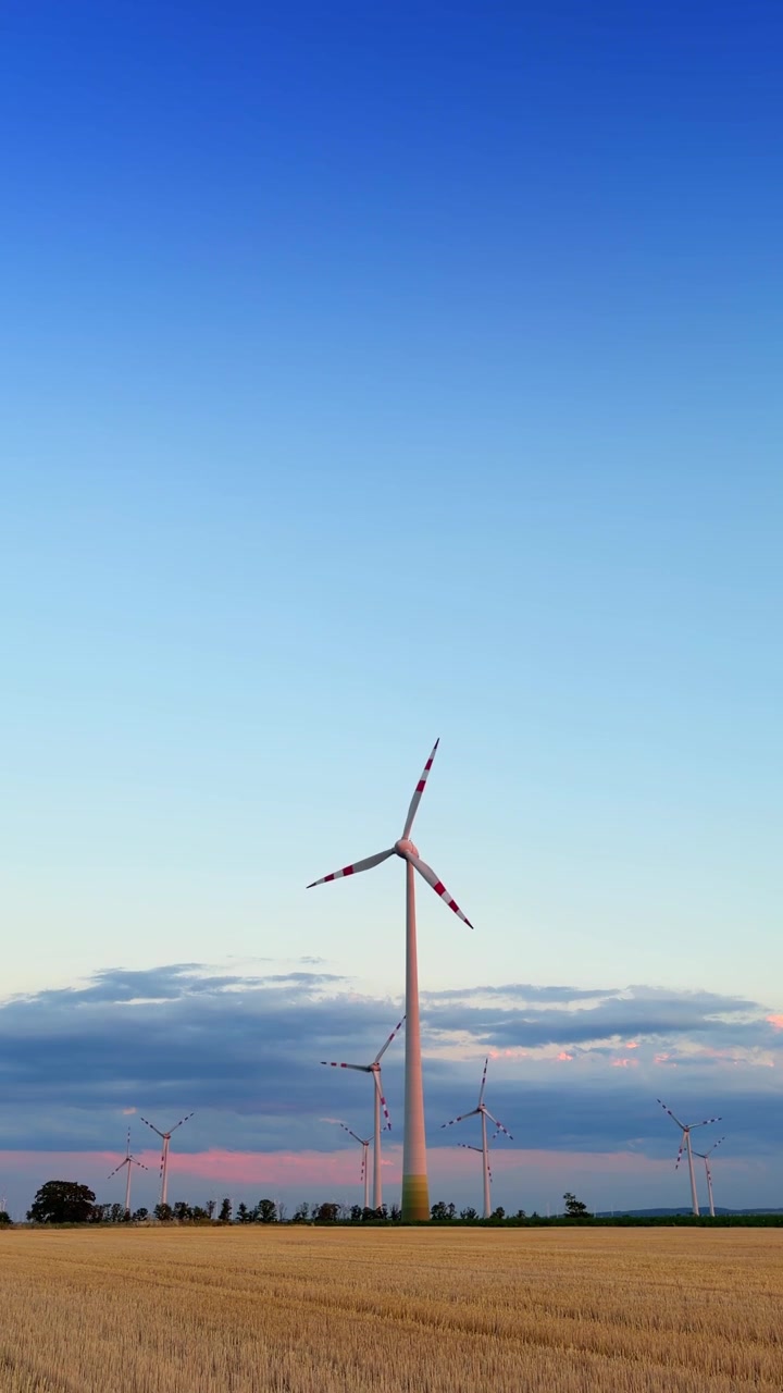 Wind turbines stand in a field at sunset. Wind turbines rotate in a wide field under a clear sky during sunset with colors changing in the background.