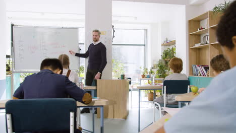 male english teacher explaining past simple to primary students on the whiteboard in classroom while pupils taking notes