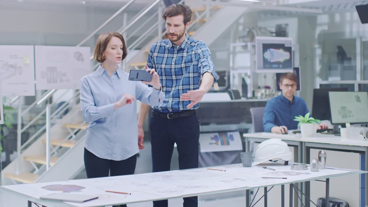 In the Industrial Engineering Facility: Female Designer with Chief Engineer Uses Smartphone for Augmented Reality App to Manipulate Engine Design Technical Drafts in 3D. High Tech Office. Mock-up