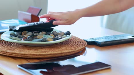 a hand reaches for and picks up a remote control from a woven placemat on a sunlit wooden table, surrounded by decorative stones and electronics