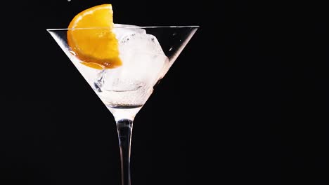 a vibrant orange slice rests on ice cubes in a clear martini glass against a dark backdrop.