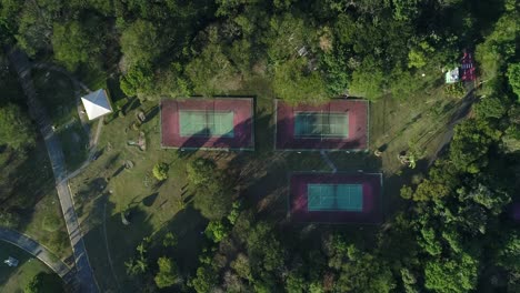 Drone-with-camera-down-descending-towards-3-tennis-courts-with-nature-around-in-public-park,-sports,-health,-leisure,-in-4K-resolution