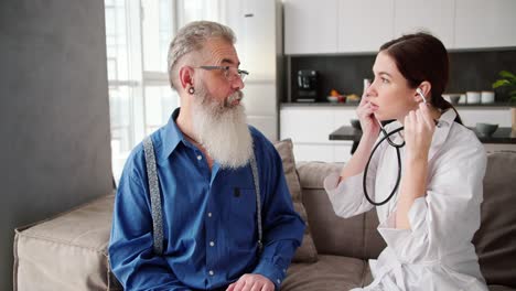 A-man-with-gray-hair-in-glasses-with-a-lush-beard-and-in-a-blue-shirt-listens-to-the-doctors-girlfriends-verdict-after-an-examination-with-a-stethoscope-in-a-modern-apartment-at-home