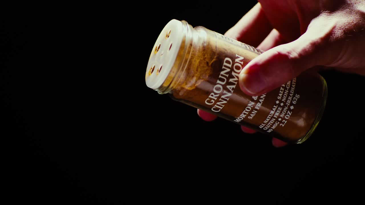 Chef's hand elegantly sprinkling Cinnamon powder against a black background. Slow motion shot with professional studio lighting