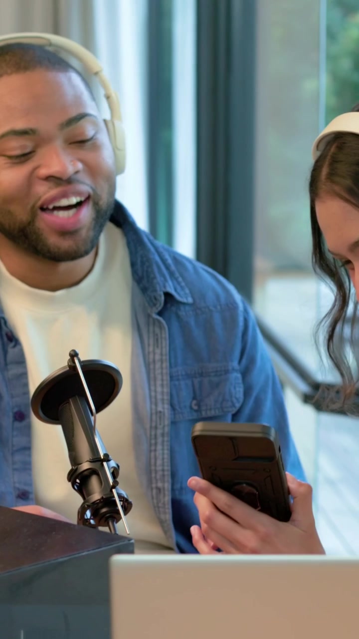 Video verticale: Donna e uomo appoggiati con le cuffie, l'uomo mostra il telefono in studio usando un microfono.