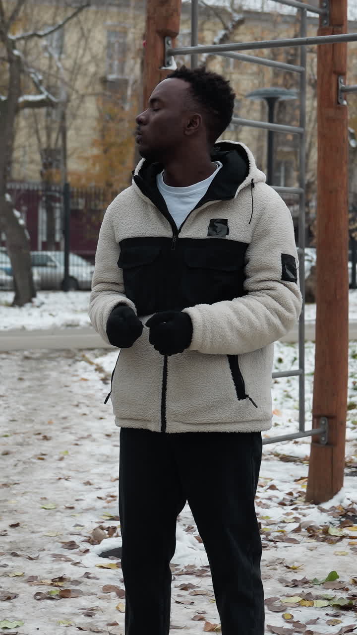 Black boy in winter jacket doing neck twist outdoors amidst workout equipment, hands joined together, background featuring moving car and parked cars covered in snow