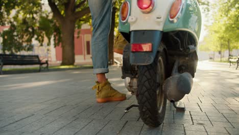 disparando de cerca, un tipo en pantalones cortos de vaqueros camina a su motocicleta verde, se sienta en ella y se aleja a lo largo de la calle con árboles y un césped en el verano