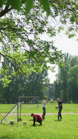 a family is doing outdoor exercise with son in red ties his shoelaces near a goalpost while his grandfather playfully hits his grandson's head with a ball