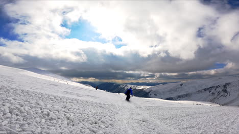following a snowboarder in black and blue outfit riding down the slope. sportsman approaches a group of people standing and falls near them. spectacular mountains and dramatic cloudscape at backdrop.