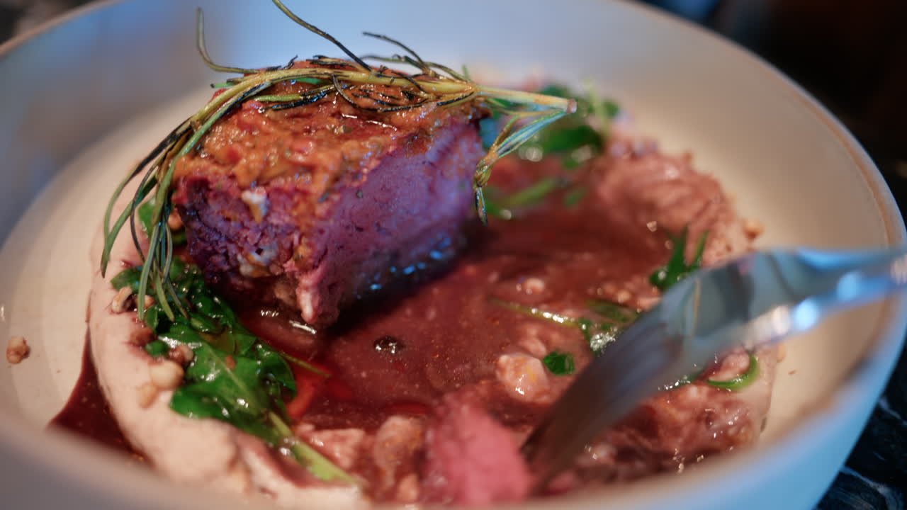 Close up of a woman's hand cutting into a gourmet meat dish served with sauce, greens, and rosemary garnish on a white plate