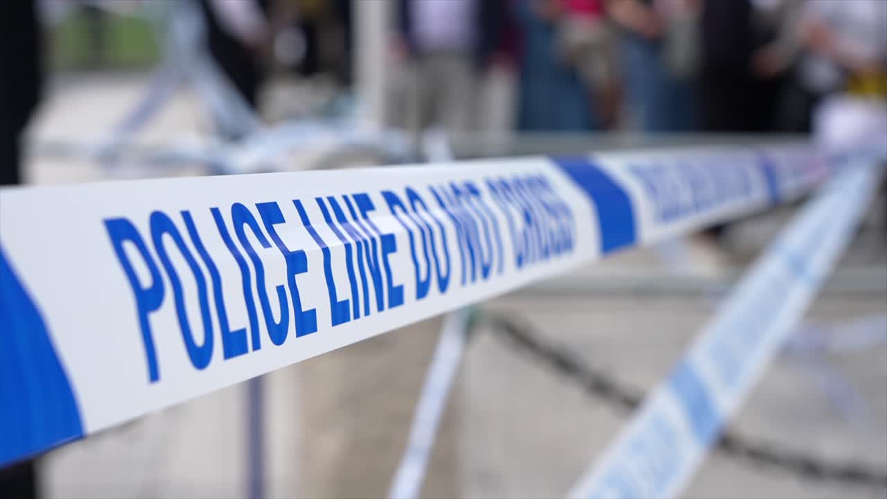 Blue and white “Police Line Do Not Cross” cordon tape flutters in the wind at an ongoing crime scene as people walk past.