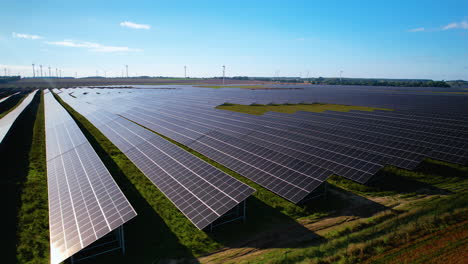 在藍天和陽光下在田野上的大型光伏太陽能發電廠的空中貨車拍攝 - 背景是風力輪發電廠