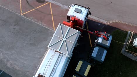 el camión de basura está recogiendo vidrio reciclado de contenedores a la orilla de la carretera, levantándolos y descargándolos en la parte trasera del camión, vista aérea superior de drones durante un día soleado, concepto de reciclaje de vidrio