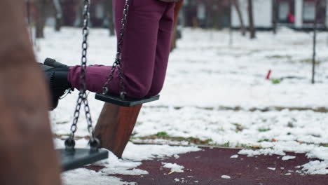 leg view of person wearing maroon trousers and black boots on swing with snow-covered ground, blurred background showing bare trees, building, and passing car