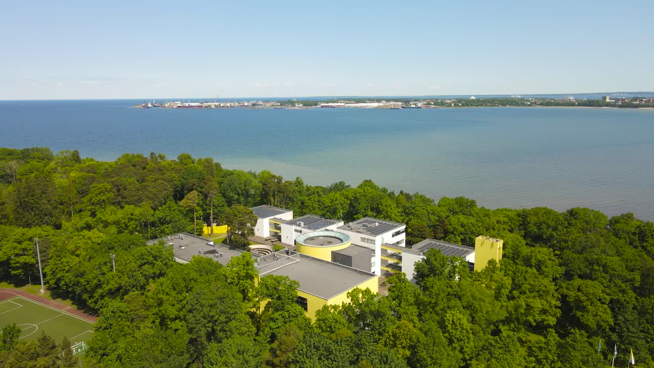 Gorgeous aerial drone footage view orbiting around Rocca al Mare highschool education building on a summer warm baltic sea shoreline in Tallinn with harbor visible on the horizon. Large green trees.