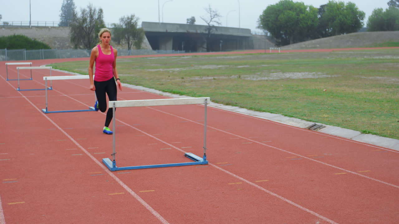 카우카스 여성 운동 선수가 4k 경주 트랙에서 장애물을 뛰어넘는 측면 뷰