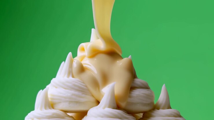 Yellow cream streams onto stacked white dessert swirls in tight close up, creating glossy folds, soft peaks, and rounded dollops against bright green studio background with fresh sweet commercial