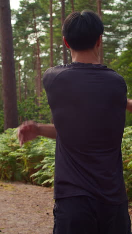 video vertical de vista trasera de un hombre calentándose y estirando los brazos antes de hacer ejercicio corriendo por el bosque 1