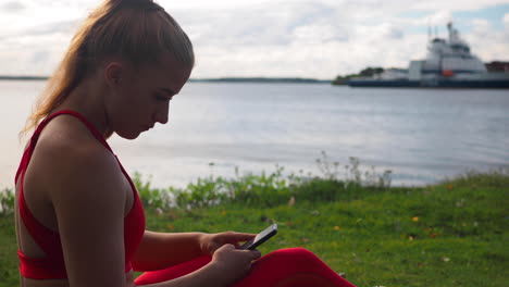 niña en ropa deportiva roja sentada en el césped en la playa y desplazándose en el teléfono inteligente