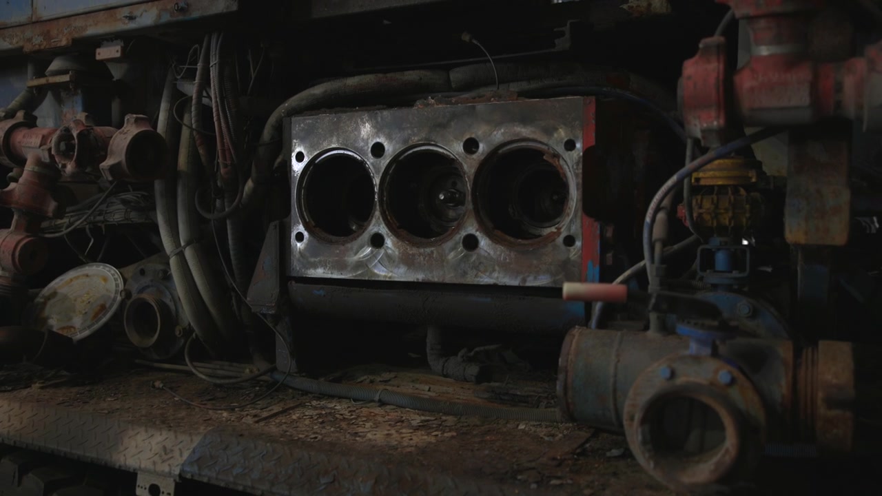 Industrial service truck midsection inside workshop bay, showing exposed pump housing, pipes, valves, hoses, dual rear wheels, rusted metal surfaces, grime, scuffed paint and heavy maintenance details
