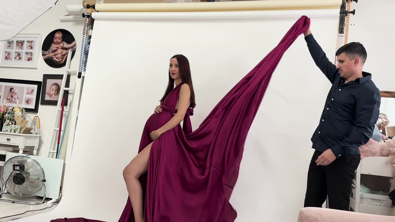 Donna incinta in uno studio che posa in un elegante abito bordeaux con un fotografo che sistema il tessuto.